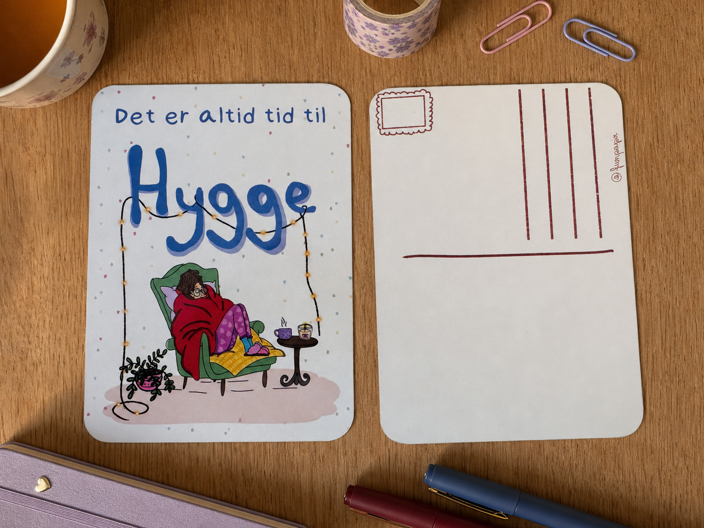 A square printed card or postcard set on a wooden desk, showing the front and back. The front has the text 'Det er altid tid til Hygge' and a cozy illustration of a person relaxing in a chair.