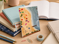 A beach-themed notebook or card propped upright on a wooden desk, surrounded by stacked notebooks, an open book, pens, paper clips, washi tape, and a planner pad. The cover illustration shows a sandy beach, blue ocean, and small colorful icons.