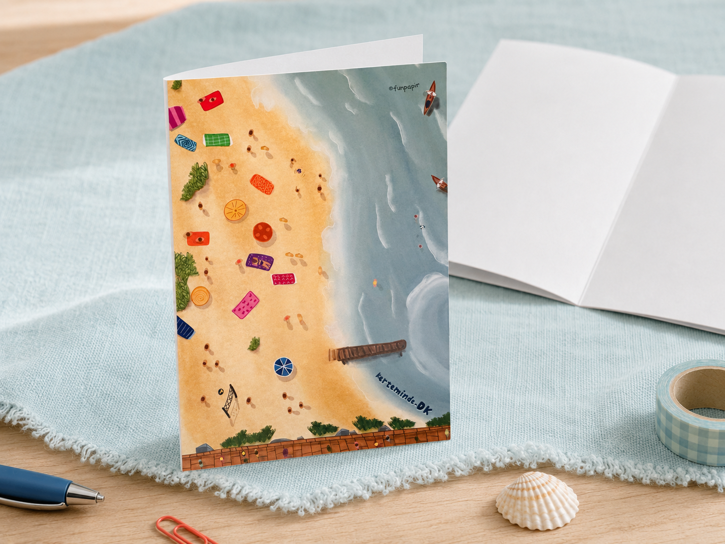 A beach-themed illustrated greeting card standing upright on a light blue fringed cloth, with an open notebook, pen, washi tape, and a seashell arranged around it on a wooden surface.