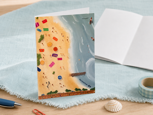 A beach-themed illustrated greeting card standing upright on a light blue fringed cloth, with an open notebook, pen, washi tape, and a seashell arranged around it on a wooden surface.