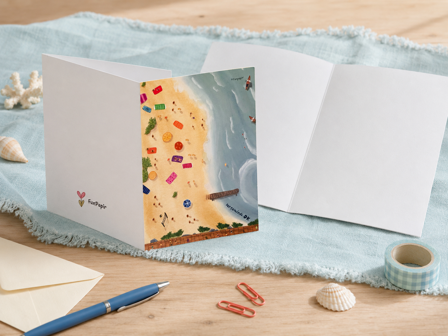 A folded greeting card displayed open on a light wood surface with a pale blue fringed cloth, shells, paper clips, a pen, and a roll of tape around it. The front of the card is white with a small colorful heart logo and the word 'together,' while the illustrated back shows a beach scene with a yellow sand area, blue water, and small multicolored icons.