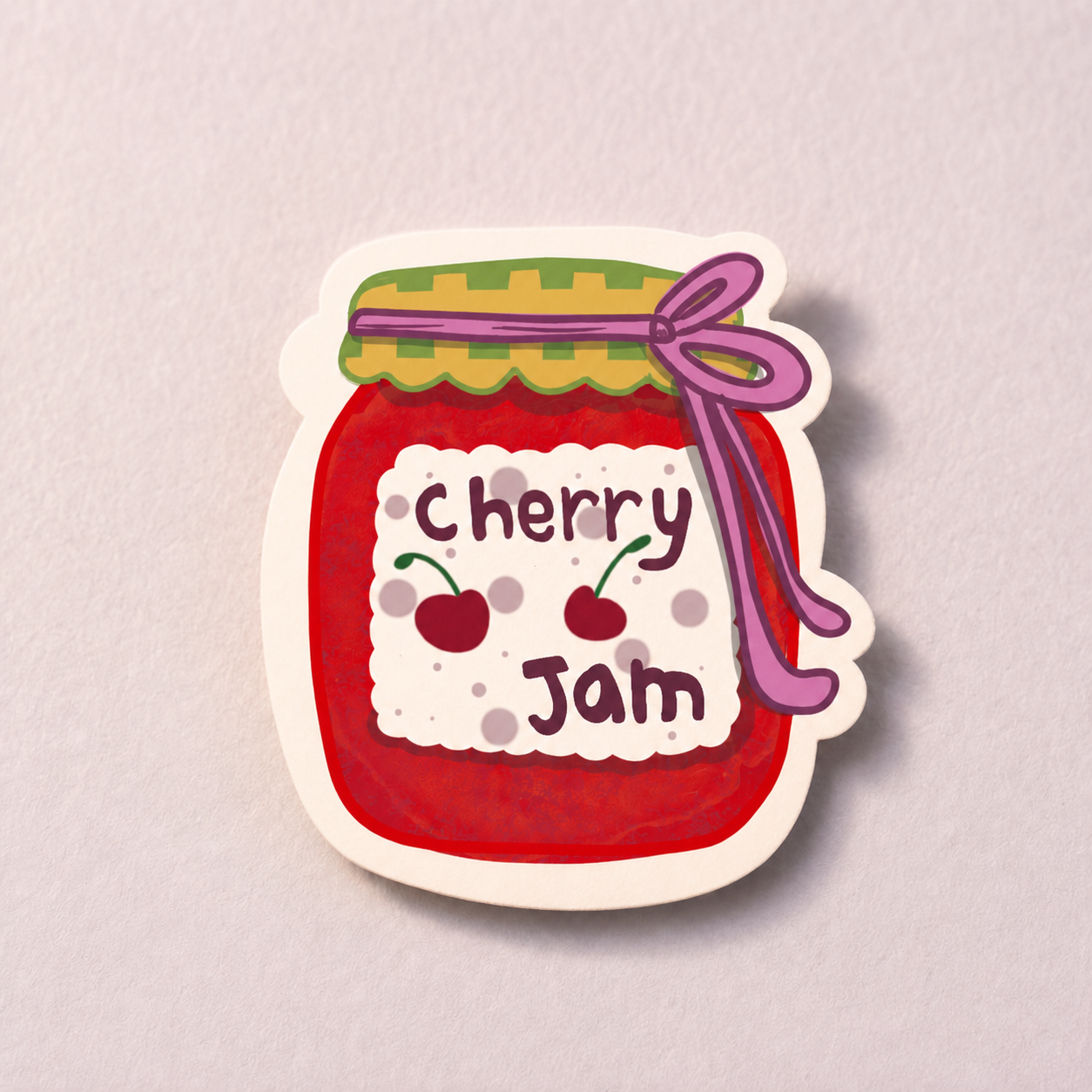 A die-cut sticker or decal shaped like a jar of cherry jam, shown on a light textured background. The sticker features a red jam-filled jar with a green and yellow patterned lid, a purple ribbon, and a white label reading 'cherry jam' with two cherries.