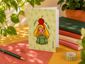 A greeting card with a cute illustration of a girl with a red cherry on her head, standing on a desk surrounded by plants, notebooks, and stationery.