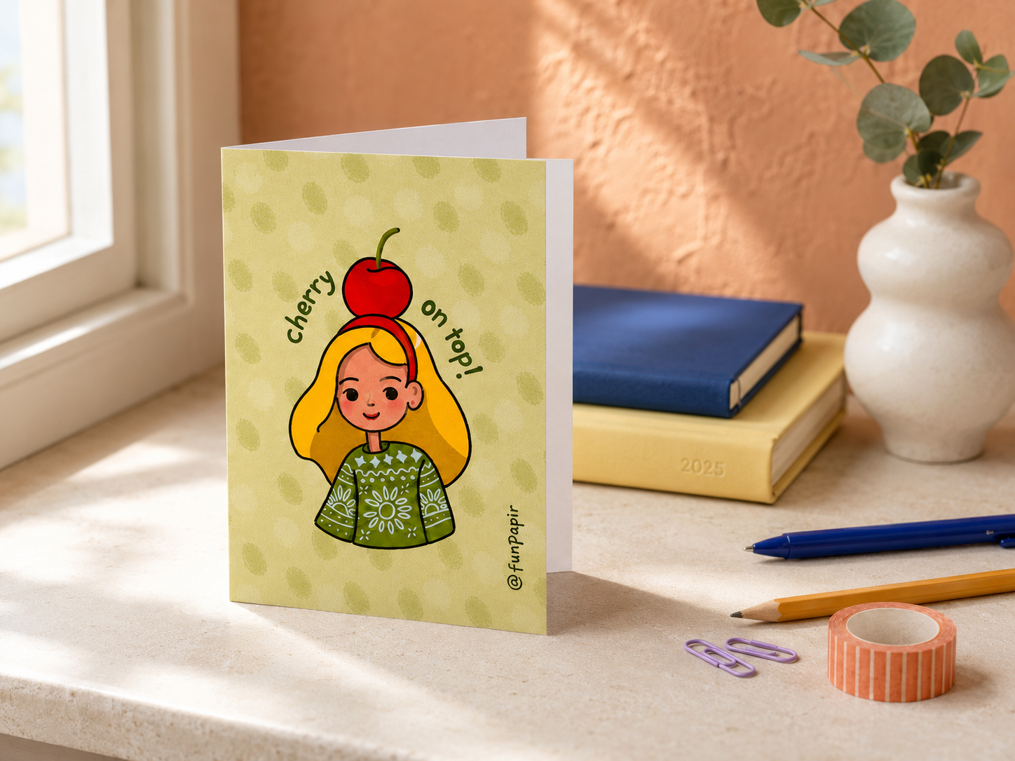 A greeting card featuring a cartoon girl with a red cherry on her head and the text 'cherry on top!' stands on a windowsill. The card has a pale yellow-green background with subtle polka dots.