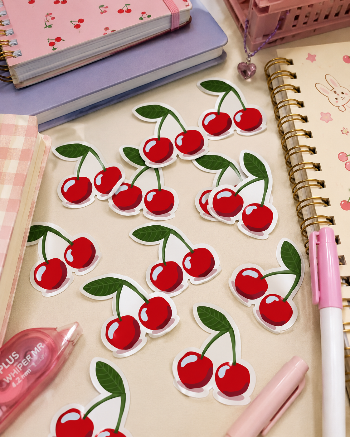 Cherry sticker set arranged on a desk with notebooks, a pink pen, and a pink correction tape dispenser. The stickers feature red cherries with green leaves and white die-cut borders.