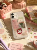 A clear phone case decorated with pastel pink and cream stickers, including a jar label reading 'Cherry Jam' and small floral photo prints, shown on a phone in a styled desk scene with notebooks, pens, and pastel accessories.