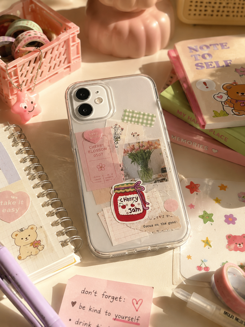 A clear phone case decorated with pastel pink and cream stickers, including a jar label reading 'Cherry Jam' and small floral photo prints, shown on a phone in a styled desk scene with notebooks, pens, and pastel accessories.