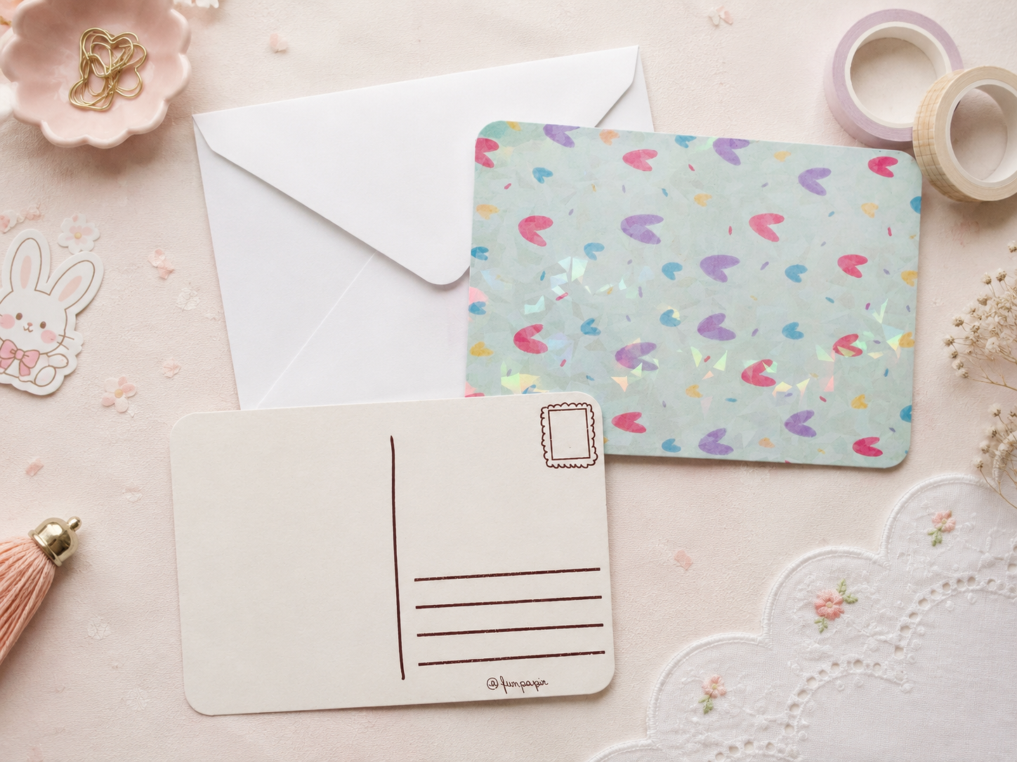 A postcard set styled on a pale pink surface, featuring a front postcard with a pastel multicolor heart pattern, a blank back postcard, a white envelope, and decorative paper accessories.