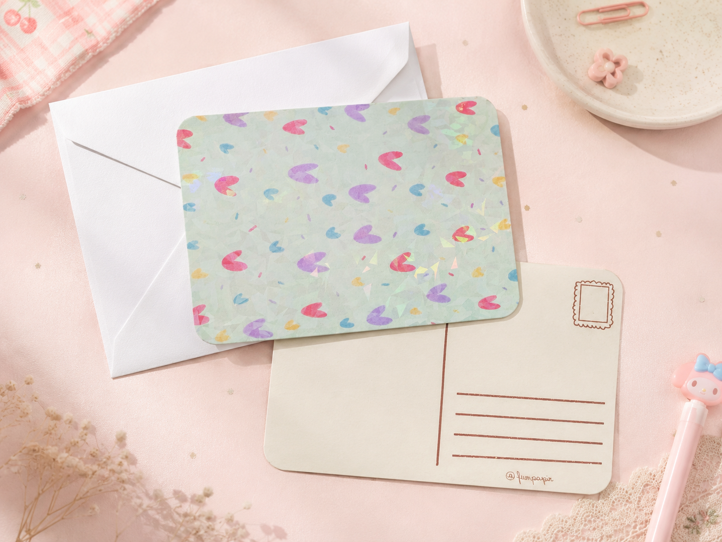 A pastel stationery flat-lay featuring a rectangular card with a mint green background and a scattered multicolor heart pattern, placed over a white envelope and a cream postcard on a soft pink surface.