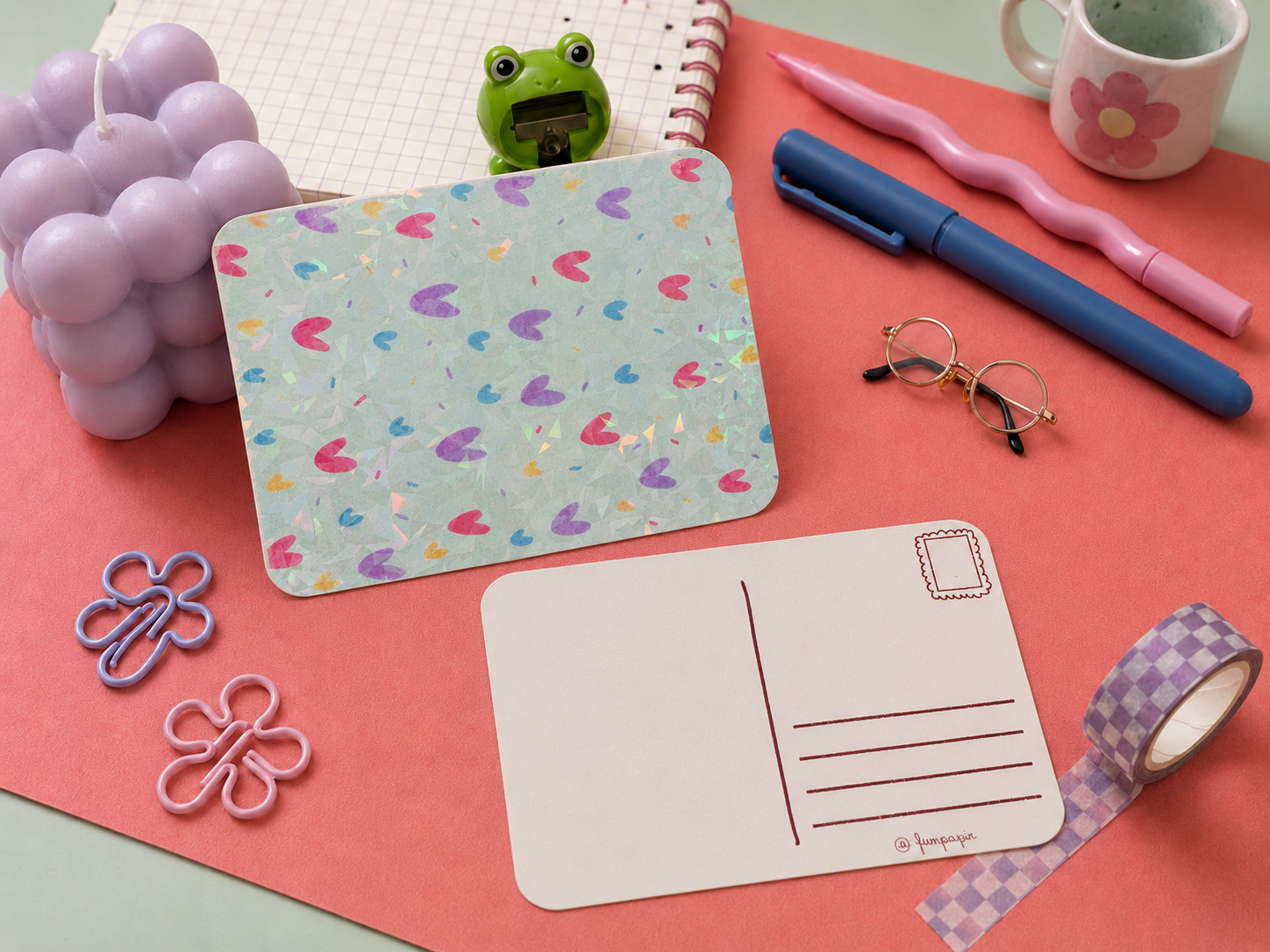 A styled flat-lay of stationery and desk accessories on a coral background, including a pastel heart-patterned mouse pad, a white postcard, pink and blue pens, a purple flower-shaped candle, a small green frog toy, eyeglasses, washi tape, and butterfly clips.