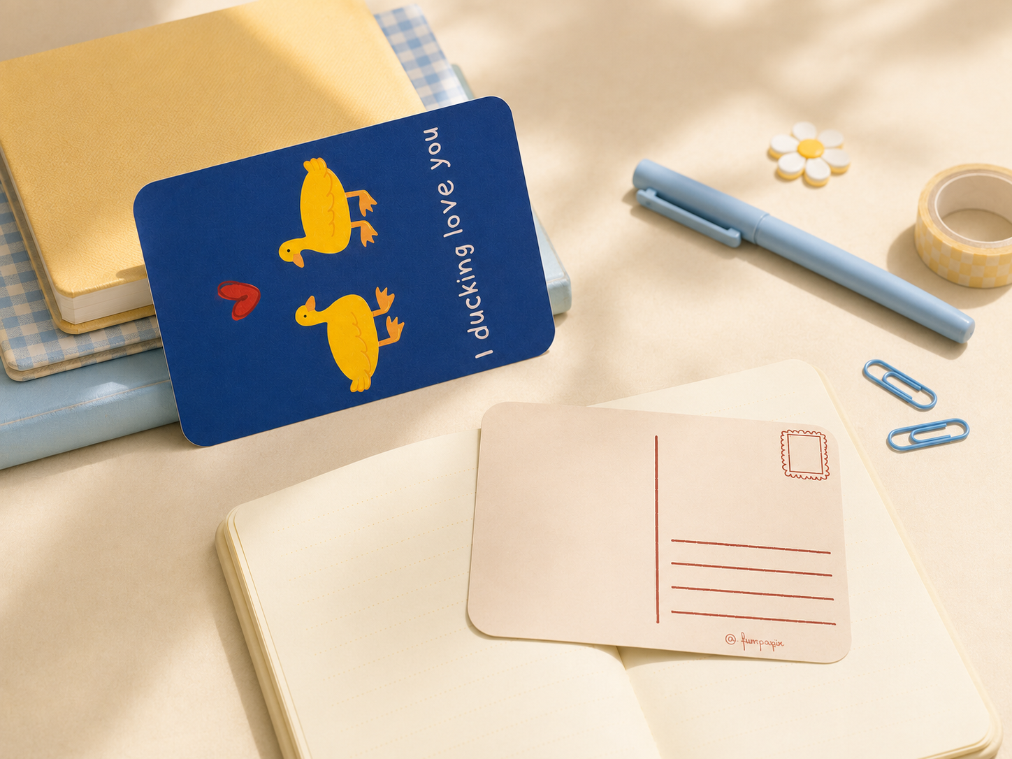 A blue postcard with yellow ducks and the text 'I ducking love, you' is displayed on a desk with notebooks, a pen, paper clips, and a postmark card.