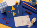 A styled flat-lay featuring a blue postcard with yellow duck illustrations and the text 'I ducking love you', alongside a blank postcard, orange pen, blue pen, red sunglasses, heart-shaped paperclips, checkered tape, a blue candle, and a rubber duck on a yellow and blue background.