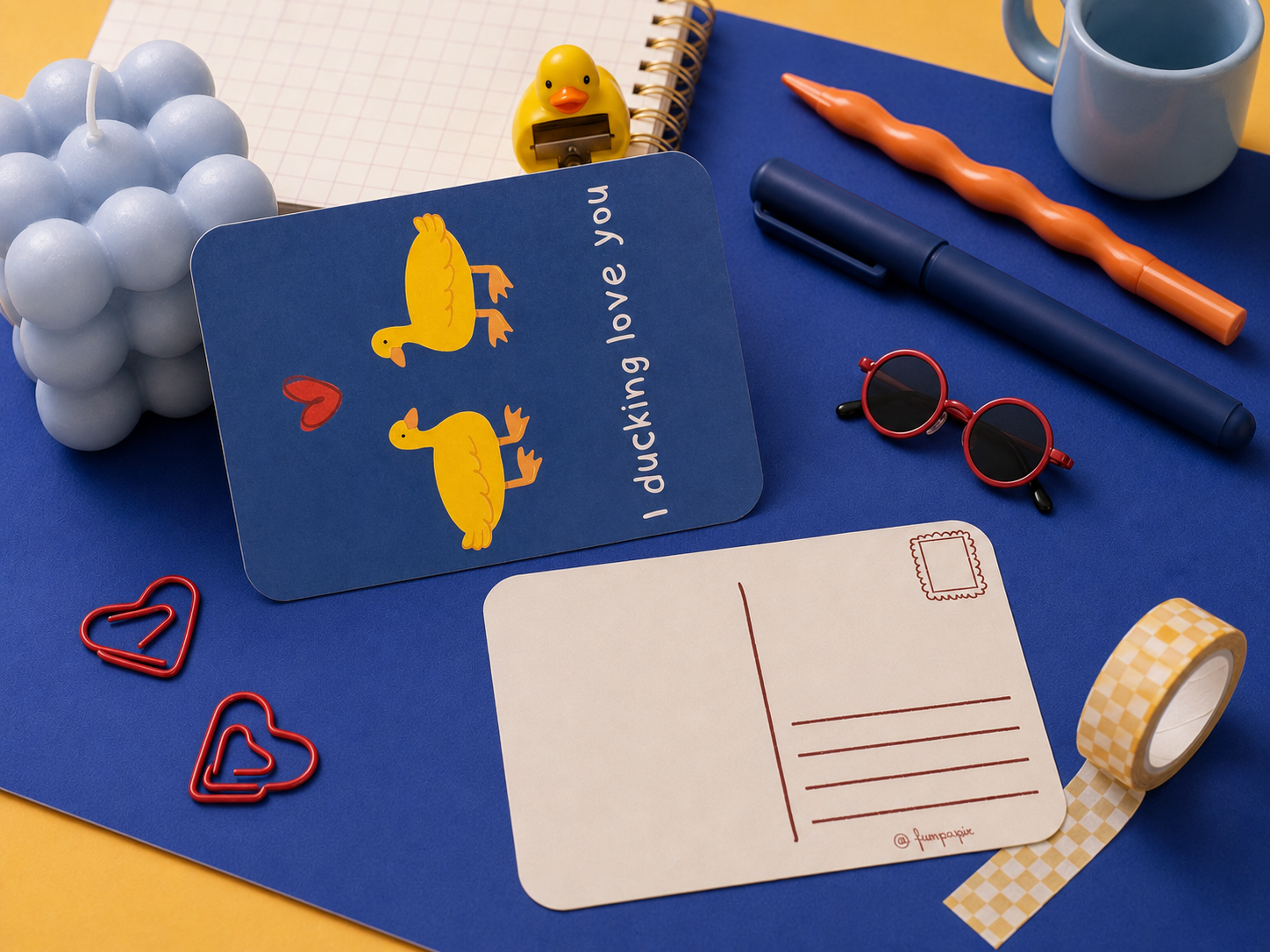 A styled flat-lay featuring a blue postcard with yellow duck illustrations and the text 'I ducking love you', alongside a blank postcard, orange pen, blue pen, red sunglasses, heart-shaped paperclips, checkered tape, a blue candle, and a rubber duck on a yellow and blue background.