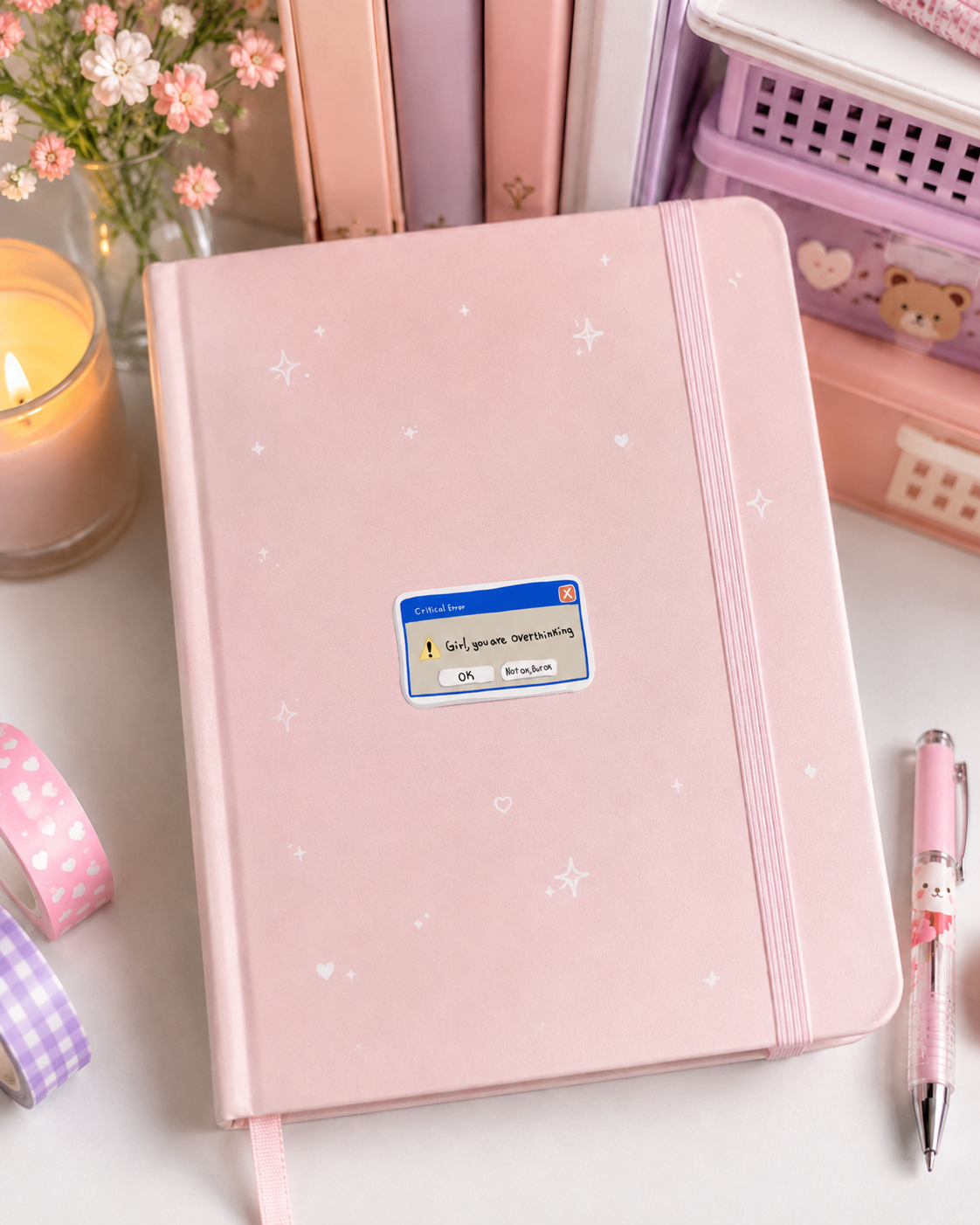 A pink hardcover notebook with a card-style cover label and small white star and moon illustrations, styled on a desk with a pen, washi tape, flowers, and stationery accessories.