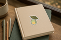 A beige hardcover notebook with a cute sticker of a green frog under a leaf umbrella, resting on a dark green notebook on a wooden desk next to a cup of coffee and pens.
