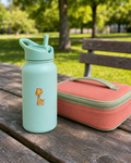 A mint green insulated water bottle with a giraffe sticker stands on a wooden picnic table next to a coral pink lunch bag with a green handle, set against a blurred park background.