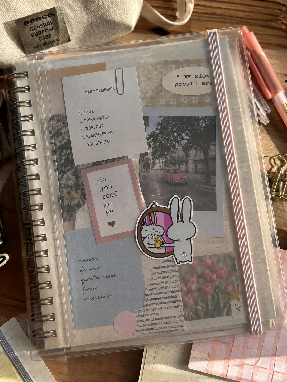 A clear plastic spiral-bound planner or notebook cover with decorative inserts, shown on a desk with pens, clips, and other stationery around it.