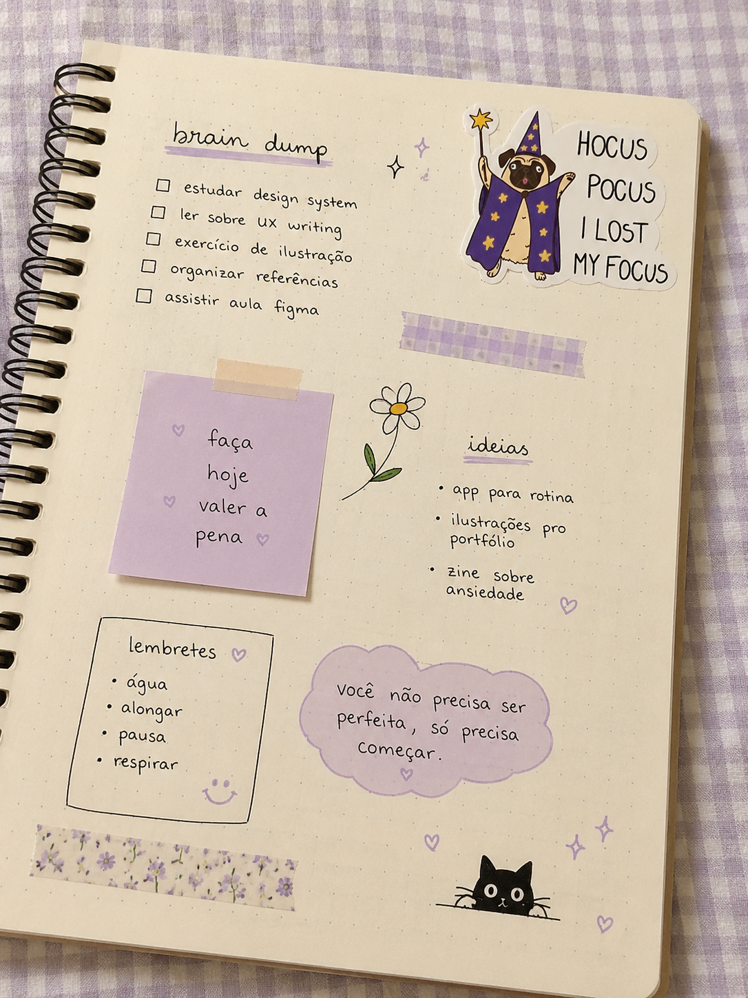 A spiral-bound notebook with Portuguese text and doodles, including a small cat illustration and decorative labels, placed on a purple-and-white checkered fabric surface.