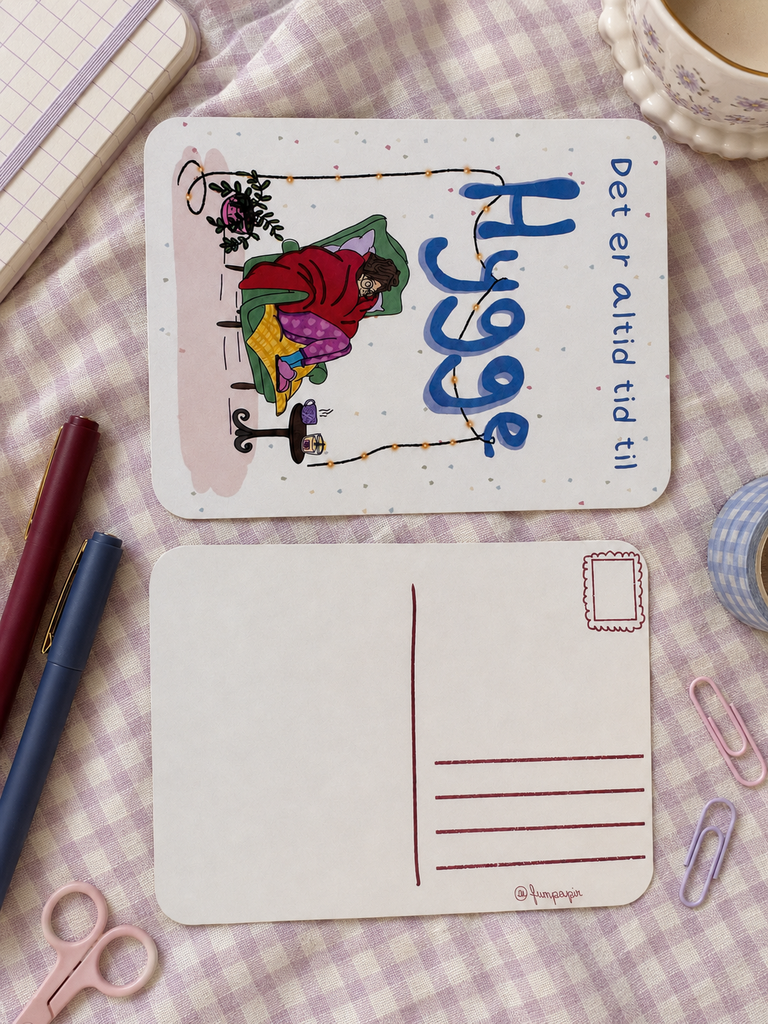 A flat-lay view of a postcard set on a lavender and white gingham fabric background. The top card features a whimsical illustration of a person in a red coat and green hat holding a string with a clock hanging from it, alongside the Swedish text 'Hjänge' and 'Det er alltid tid till'.