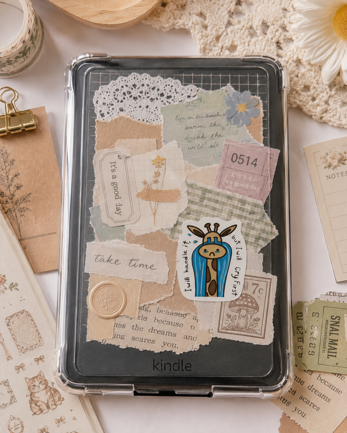 A Kindle device with a decorative vintage-style collage cover, shown from above on a styled flat-lay background with paper clips, lace, and retro paper pieces.