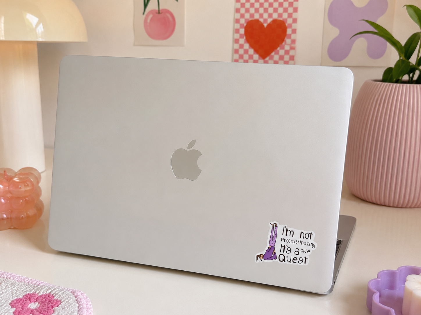 A silver Apple laptop sits on a desk in a styled room setting, featuring a custom sticker on the bottom right corner that reads 'I'm not overanalyzing it's Quest' with a purple giraffe illustration. The background includes a pink ribbed planter, a cream-colored lamp, and decorative wall art.