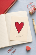 A cream-colored greeting card with a red heart and the word 'love' written in cursive, placed on an open notebook. The scene is styled with a red pen, paper clips, a spool of twine, and a small red heart on a light blue surface.