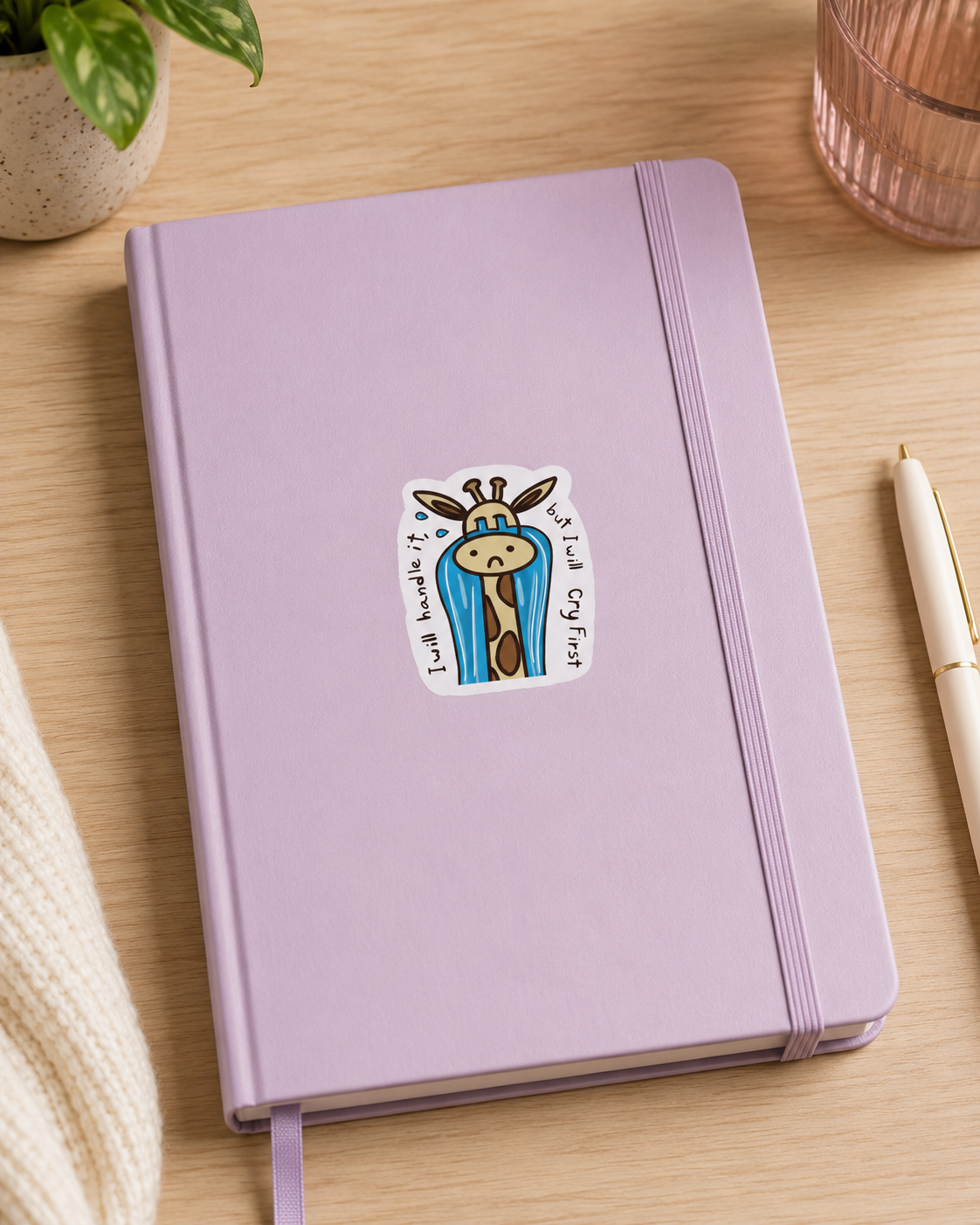 A lavender purple hardcover notebook with an elastic closure band, featuring a cartoon giraffe sticker on the front, styled on a light wood desk with a pen, plant, and glass.