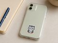A clear phone case on a light sage green iPhone, featuring a cute cartoon owl sticker on the back. The phone case is displayed on a beige desk next to a blue pen and notebook.
