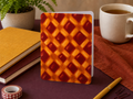 A card with a pie pattern standing upright on a desk, surrounded by a stack of notebooks, a plant, a mug of coffee, a pen, and washi tape.