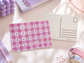 A pink and white gingham postcard holder with small red heart patterns, shown next to a blank postcard on a styled desk with pastel stationery props.
