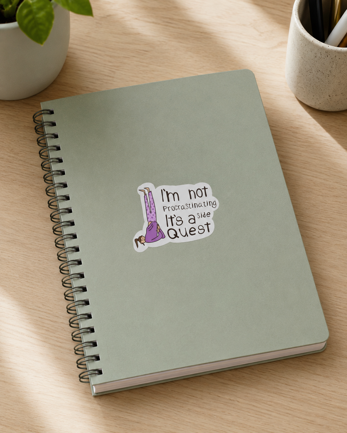 A sage green spiral-bound notebook with a humorous sticker that reads 'I'm not Practicing It's a Real Quest' featuring a purple potion bottle, resting on a light wood desk with a plant and cup in the background.