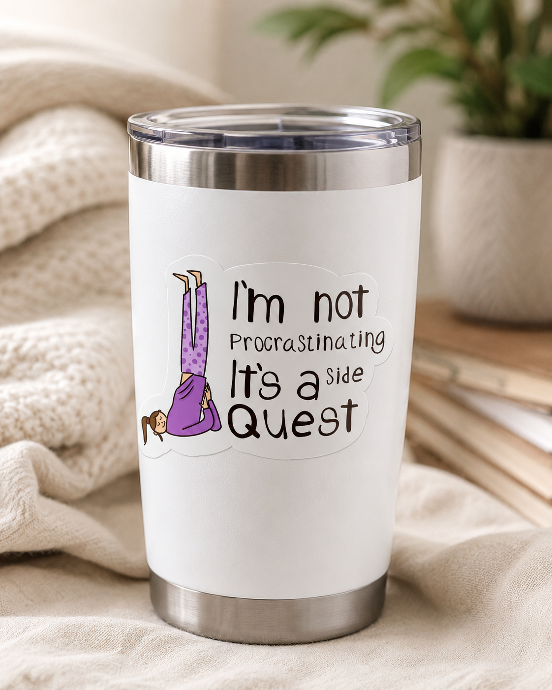 A white insulated travel tumbler with a clear lid, featuring a printed graphic of a person doing a headstand in purple and black text that reads, 'I'm not Procrastinating It's a Quest.' The tumbler is shown on a soft beige fabric surface with a cozy indoor background and a blurred plant.
