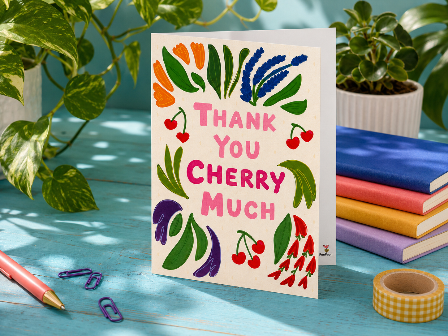 A greeting card with a colorful botanical illustration and the text 'THANK YOU CHERRY MUCH' in pink, standing on a painted blue wooden table. The card is styled with books, washi tape, paper clips, and potted plants.