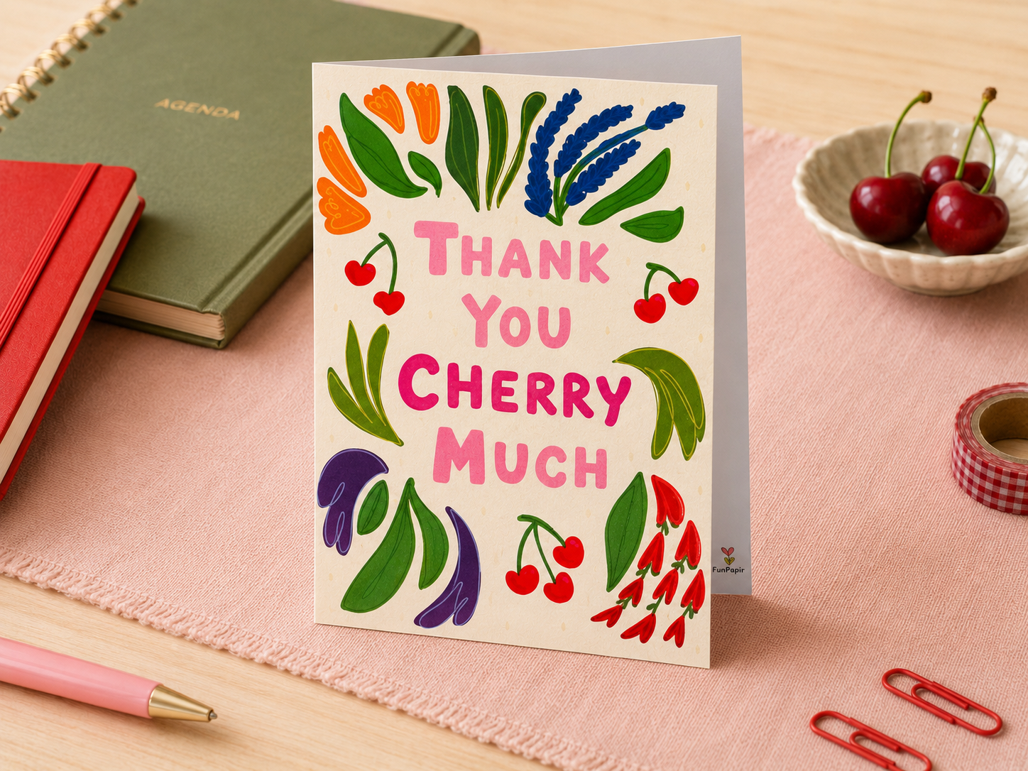 A greeting card standing upright on a pink cloth, surrounded by stationery props including a notebook, pen, cherries in a bowl, and paper clips. The card reads “THANK YOU CHERRY MUCH” with colorful cherry and leaf illustrations.