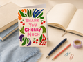 A square greeting card with a cream background and colorful hand-drawn cherry and leaf illustrations, standing upright on a desk with an open book, notebook, pen, pencil, paper clips, and washi tape around it. The card reads “THANK YOU CHERRY MUCH.