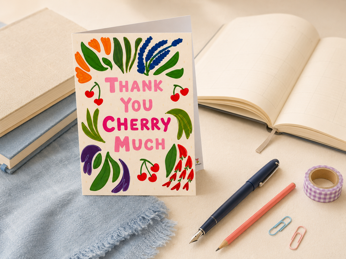 A square greeting card with a cream background and colorful hand-drawn cherry and leaf illustrations, standing upright on a desk with an open book, notebook, pen, pencil, paper clips, and washi tape around it. The card reads “THANK YOU CHERRY MUCH.