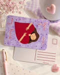 A rectangular greeting card or note with rounded corners lies on a light surface. The front shows a lavender background with small heart motifs, a central illustration of a woman in a red dress, and the words 'my heart you are' in pink script.