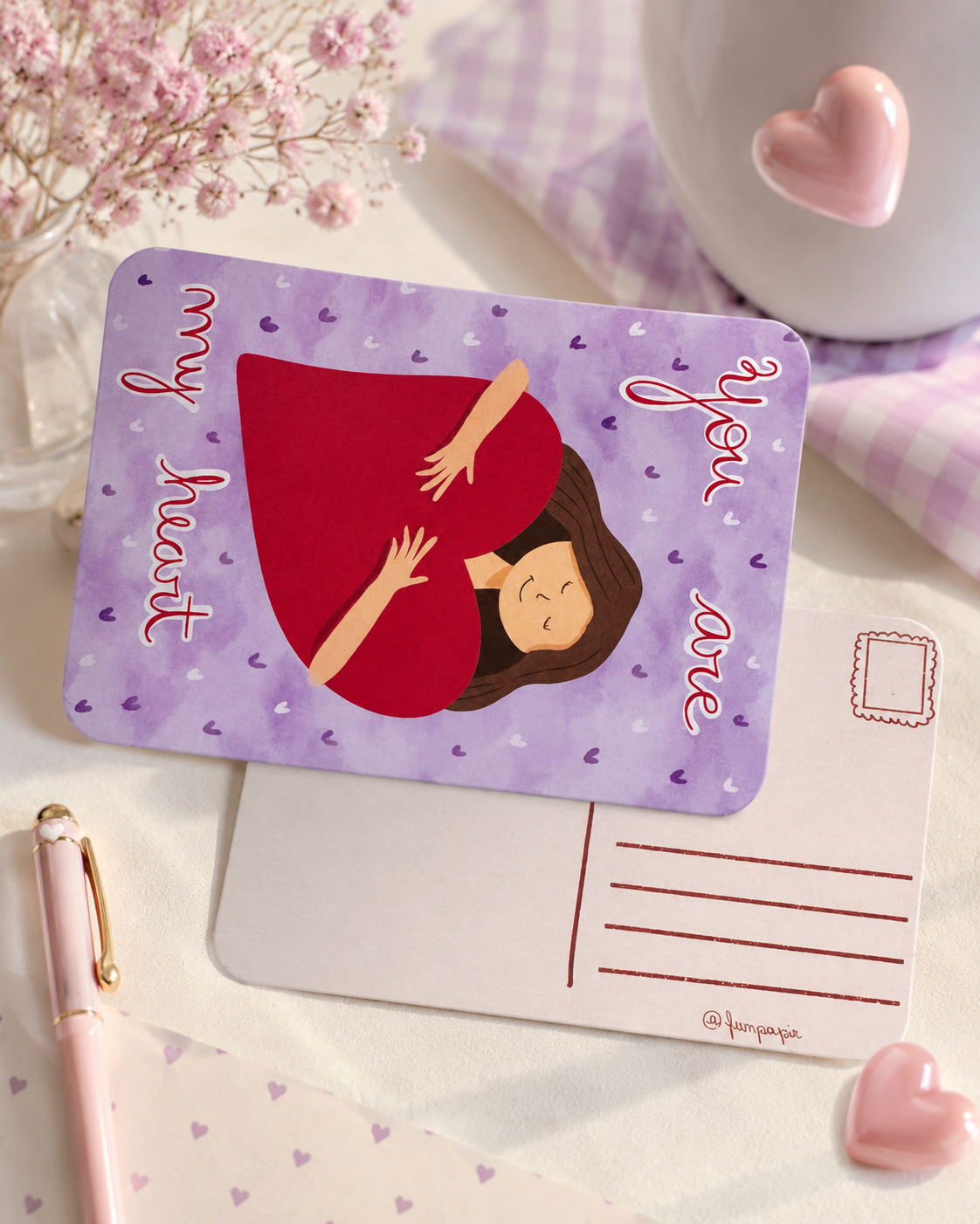 A rectangular greeting card or note with rounded corners lies on a light surface. The front shows a lavender background with small heart motifs, a central illustration of a woman in a red dress, and the words 'my heart you are' in pink script.