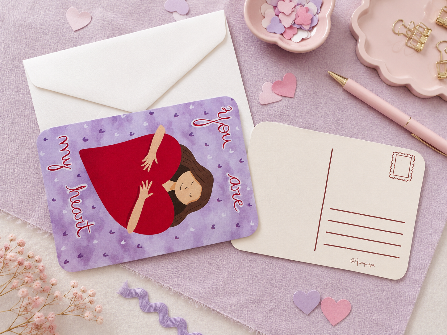 A flat-lay of a postcard and matching envelope on a lavender fabric background. The postcard features a purple background with small heart motifs, a red heart-shaped silhouette of a pregnant woman, and the words 'my heart' and 'you are' in pink script.
