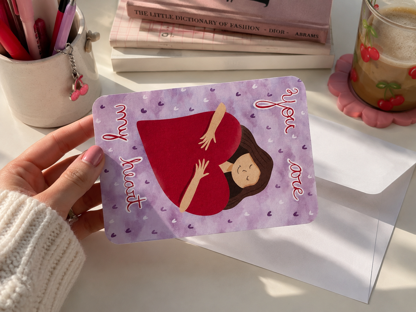 A hand holds a purple greeting card featuring an illustration of a pregnant woman in a red belly, with the text 'my heart' and 'you are'. The card is shown in a styled desk setting with a white envelope, pens, books, and a cherry-patterned candle.