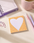 A square sticky note pad with a large white heart on a pastel yellow background with tiny heart patterns, styled on a desk with a pink notebook, paperclips, pens, and a mug.
