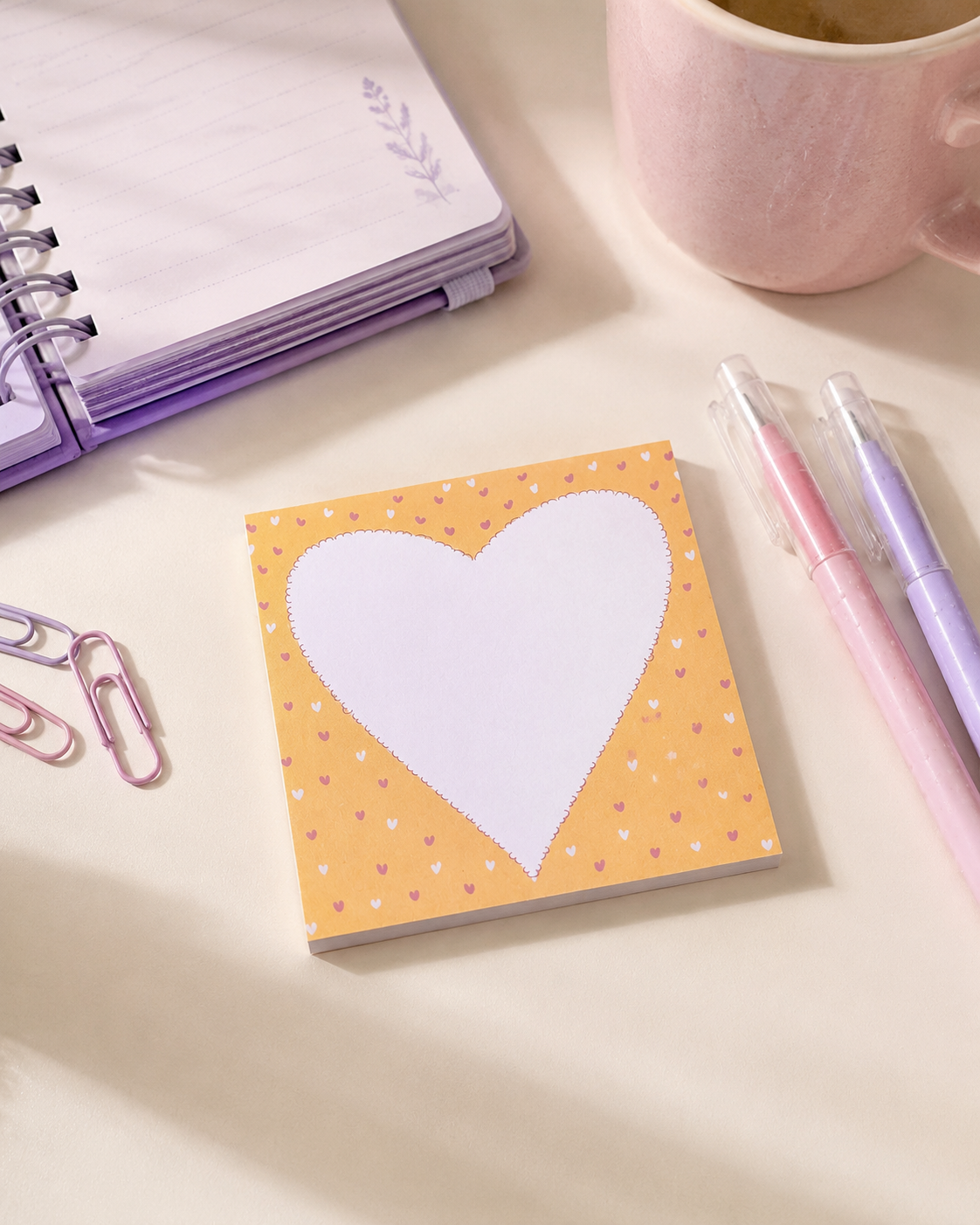 A square sticky note pad with a large white heart on a pastel yellow background with tiny heart patterns, styled on a desk with a pink notebook, paperclips, pens, and a mug.